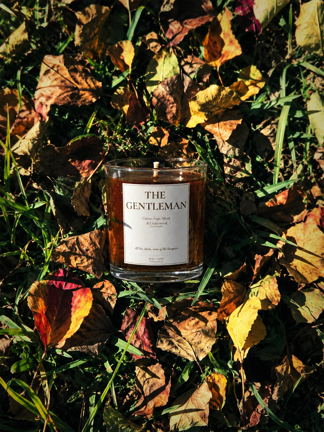 A glass jar candle labeled 'THE GENTLEMAN' sits outdoors on a bed of dry autumn leaves and grass in warm sunlight.